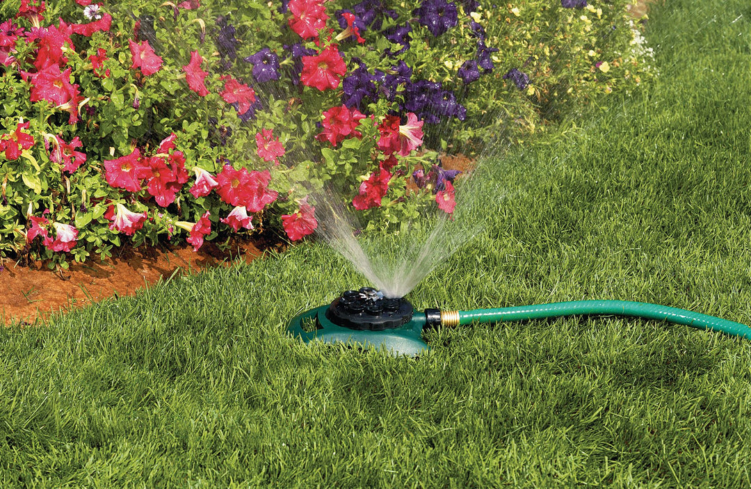 Green and black hose end 8-pattern plastic turret sprinkler spraying water on grass and flowers.